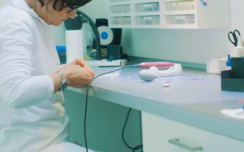 Mitarbeiterin in weißem Kittel arbeitet an einem Hörgerät auf einem Arbeitstisch.
