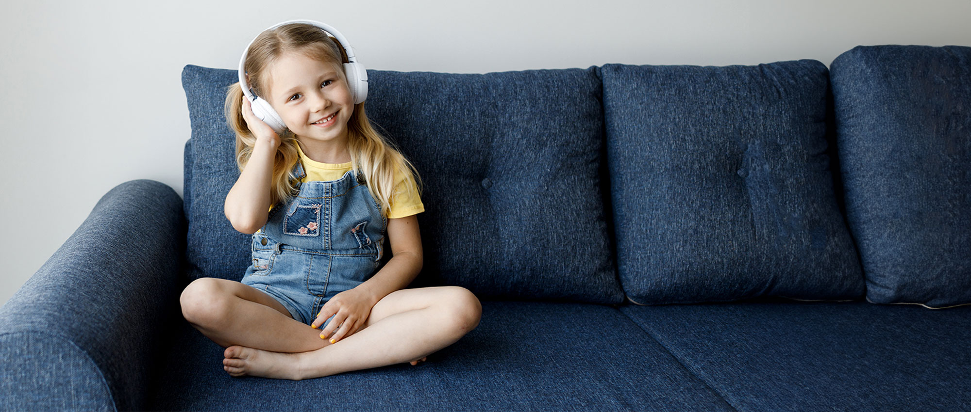 Ein Mädchen sitzt fröhlich auf einem blauen Sofa und trägt weiße Kopfhörer. Durch regelmäßige Hörtests für Schulkinder können potenzielle Hörprobleme frühzeitig erkannt werden, um die Hörgesundheit zu fördern. Ein Mädchen sitzt fröhlich auf einem blauen Sofa und trägt weiße Kopfhörer. Durch regelmäßige Hörtests für Schulkinder können potenzielle Hörprobleme frühzeitig erkannt werden, um die Hörgesundheit zu fördern.