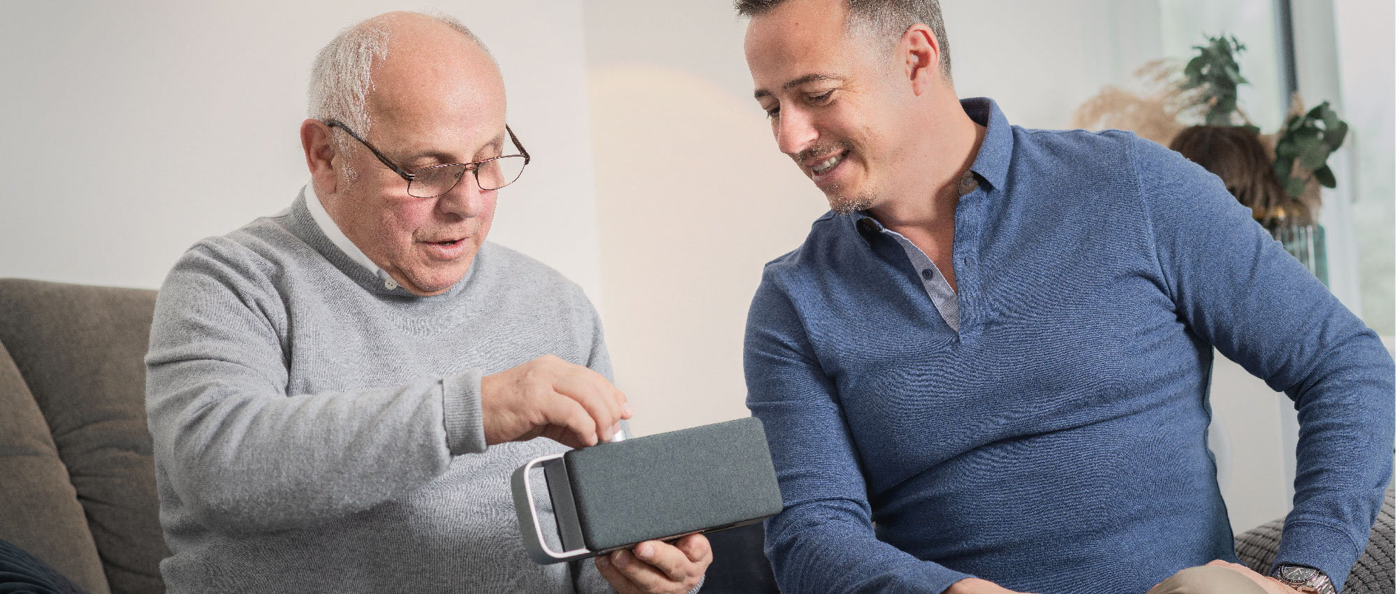 Ein älterer Mann zeigt einem jüngeren Mann den OSKAR TV-Sprachverstärker. OSKAR verbessert das Sprachverständnis bei Fernsehsendungen. Ein älterer Mann zeigt einem jüngeren Mann den OSKAR TV-Sprachverstärker. OSKAR verbessert das Sprachverständnis bei Fernsehsendungen.